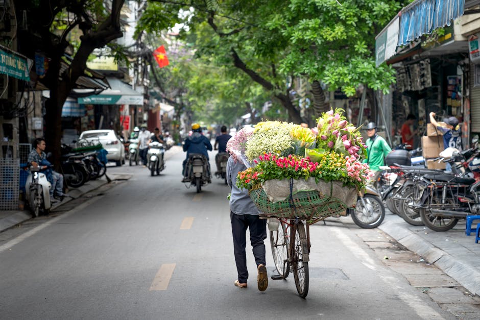 A flower seller on a busy city street pushes a bicycle loaded with a large, colorful floral arrangement. The arrangement comprises pink, white, yellow, and green flowers, including roses, hydrangeas, 