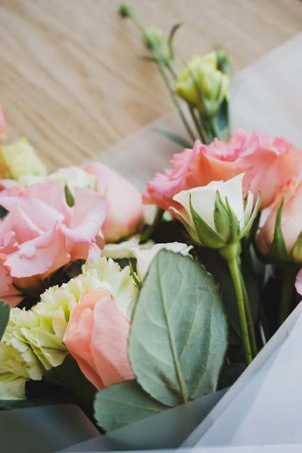 A close-up photograph of a floral bouquet featuring soft pink and cream lisianthus flowers with delicate, ruffled petals and green buds, arranged with light green, oval-shaped leaves. The flowers are 