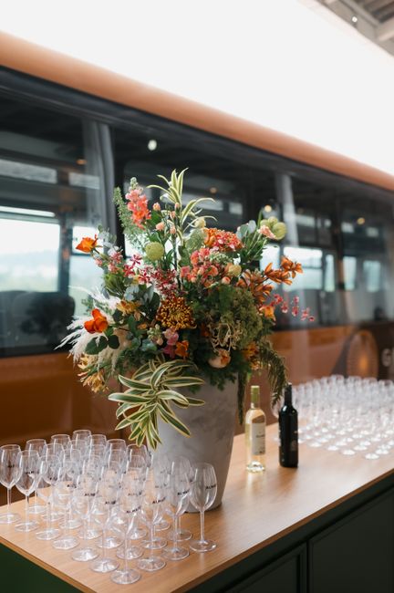 A tall, vibrant floral arrangement in a gray ceramic vase features a mix of colorful flowers including orange and pink blooms, complemented by green foliage and variegated leaves with cream and green 