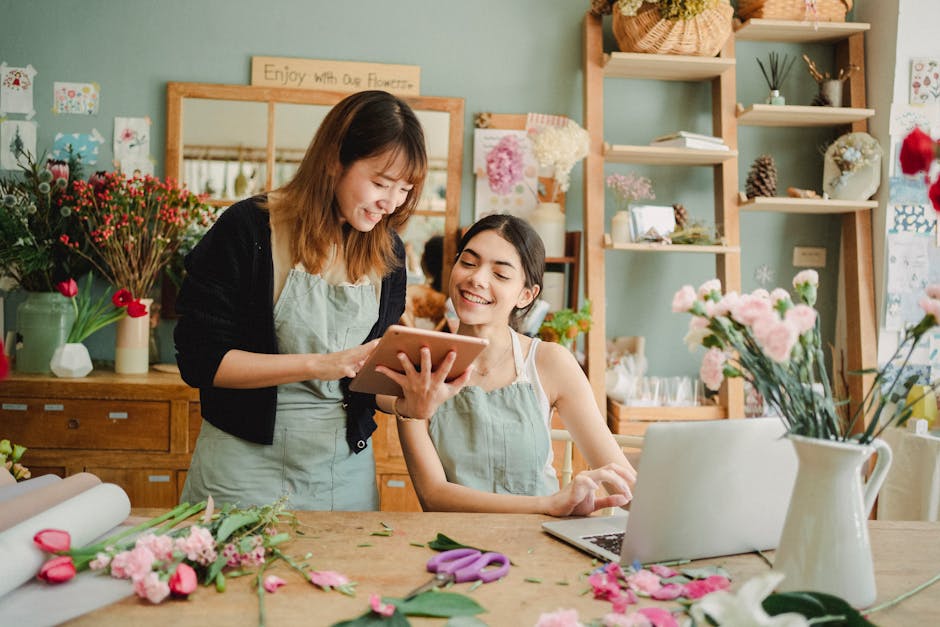 Two women working together in a floral shop, one holding a tablet and the other smiling, with various fresh flowers and floral arrangement tools spread on the wooden table in front of them. The backgr
